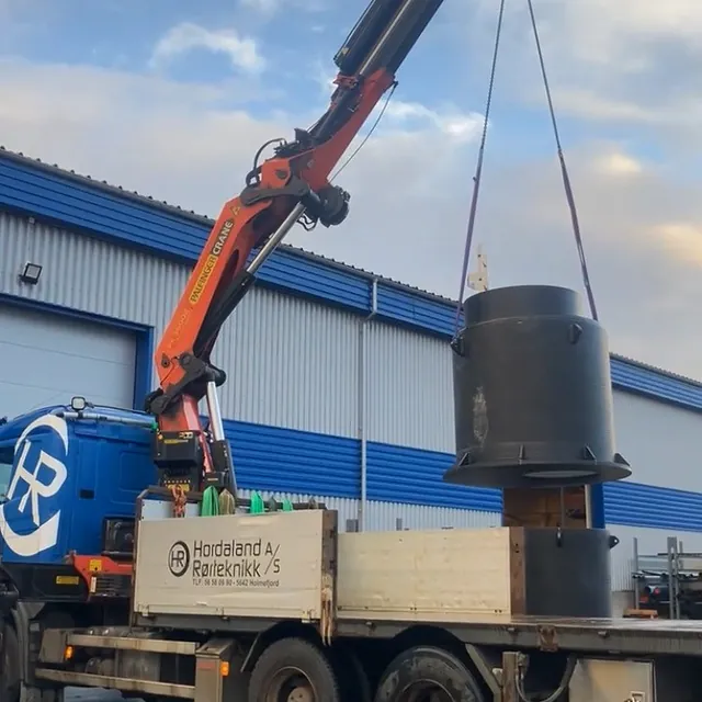 Lastebil med kran løfter stor svart kum foran industribygg.