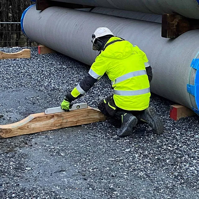 Arbeider i gul refleksjakke bruker vater ved store betongrør.