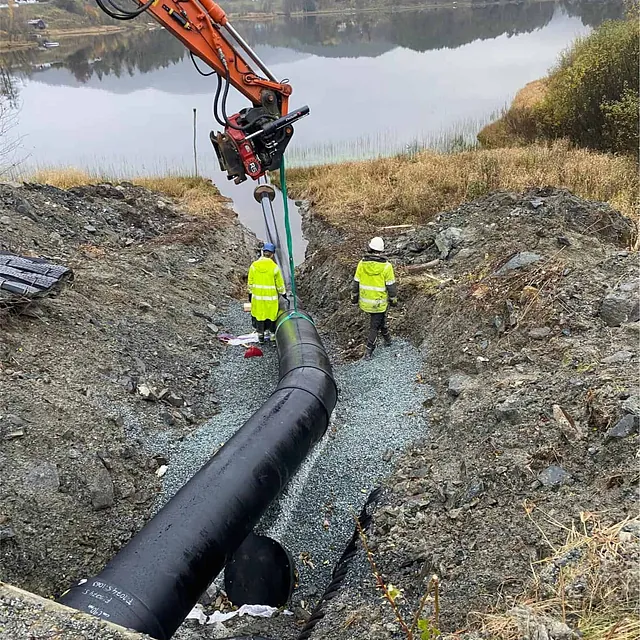 Gravemaskin løfter svart rør ned i grøft ved vann, to arbeidere følger med.