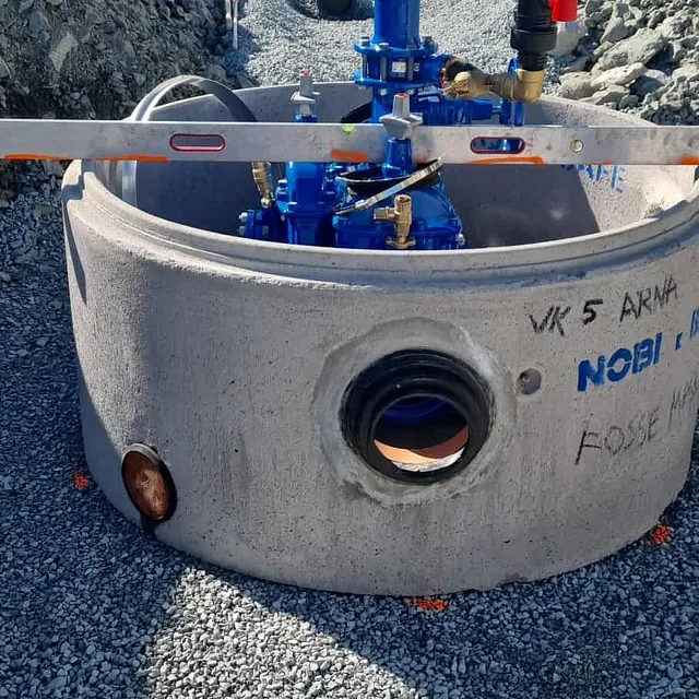 Betongkum med blå ventiler og rørarbeid i grøft omgitt av fjell.